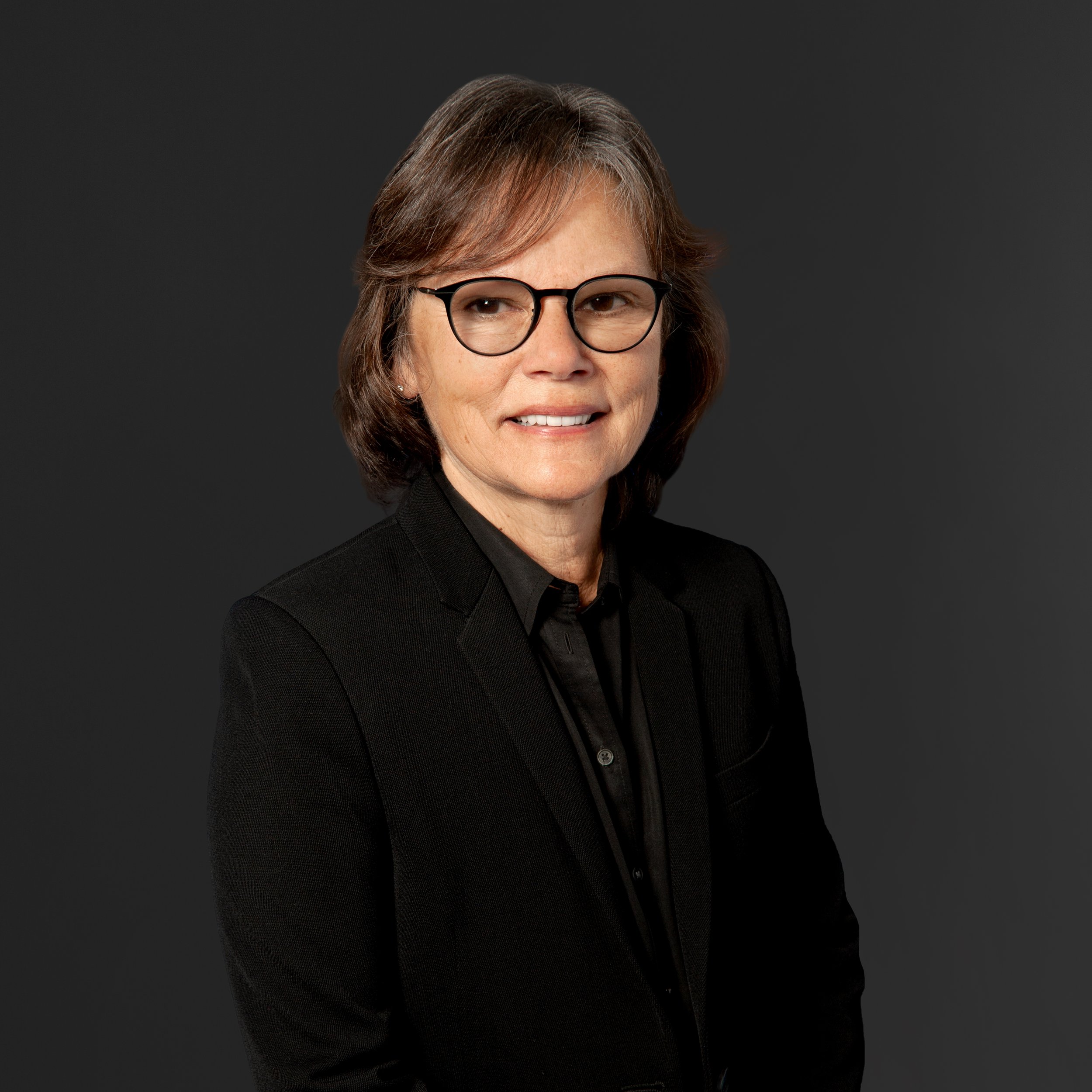 Portrait of a middle-aged woman with shoulder-length brown hair, wearing glasses, a black blazer, and a black shirt, against a dark background.