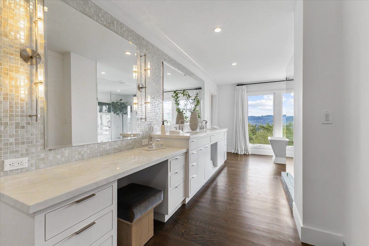 Bright modern bathroom or dressing area with white cabinetry, large mirror, mosaic tile wall, and a view through a window of trees and hills outside.