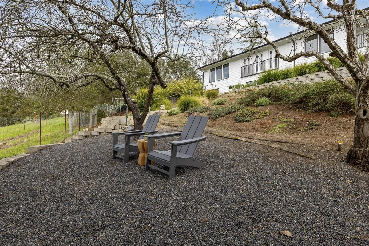 Two black Adirondack chairs and a small wooden side table on a gravel patio with trees and bushes in the background, overlooking a white house with large windows.