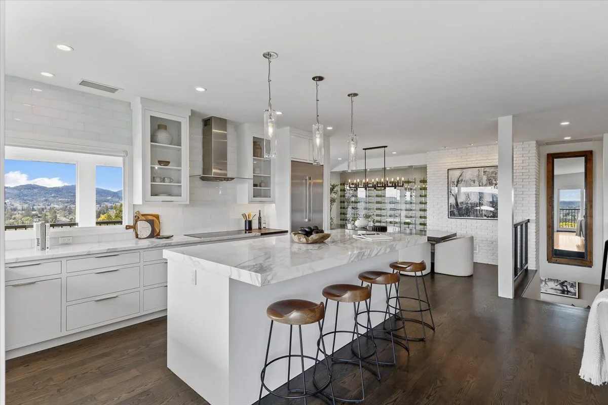 Modern kitchen with white cabinets, marble island, four wooden bar stools, pendant lights, window view of mountains and trees, white brick wall, and contemporary decor.