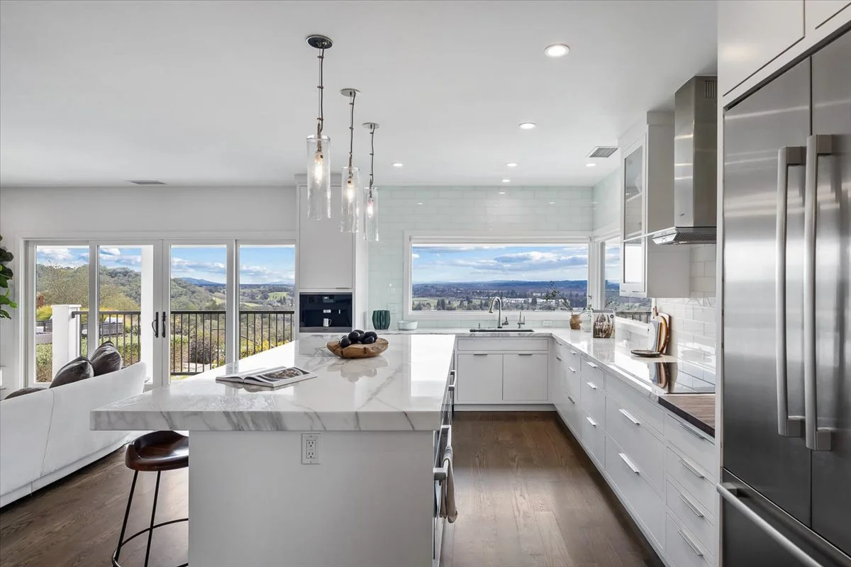 Modern white kitchen with marble island, stainless steel appliances, and large windows showing a scenic outdoor view.