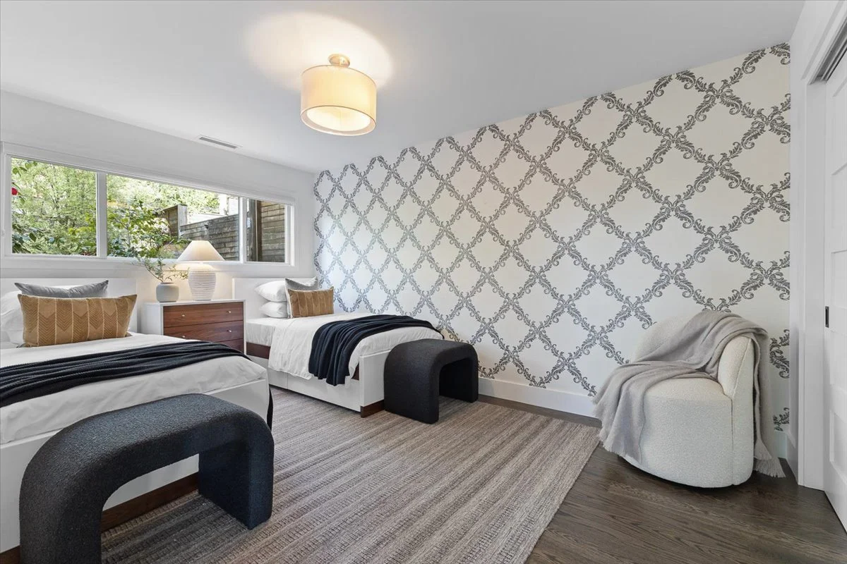 A bedroom with two twin beds, a window with a view of greenery, a nightstand with a lamp, and a patterned accent wall. There is a small armchair with a blanket draped over it and modern black stools at the foot of the beds.