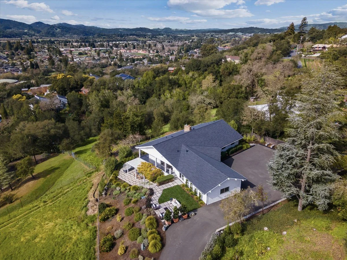 Aerial view of a suburban house with a blue roof, surrounded by trees, a driveway, and a landscaped garden in a hilly area with a distant town and mountains in the background.