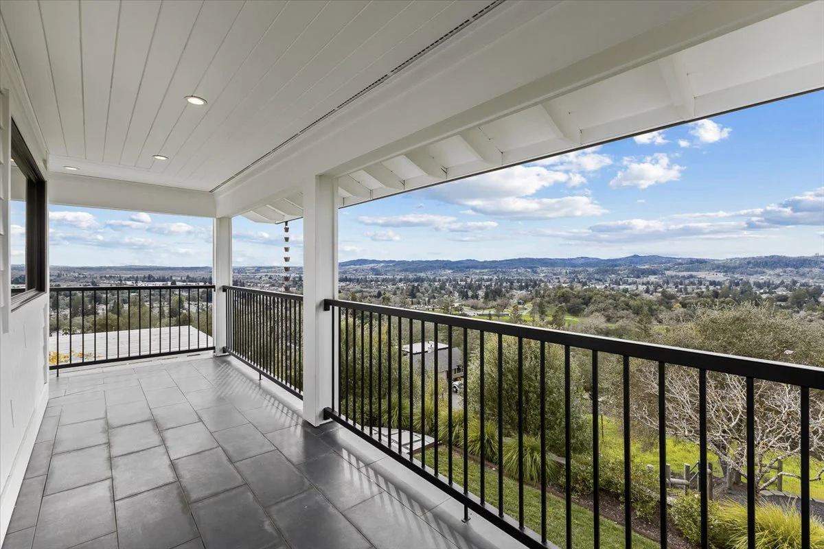 Balcony with black railing overlooking a suburban landscape with green trees, houses, and distant hills under a partly cloudy sky.