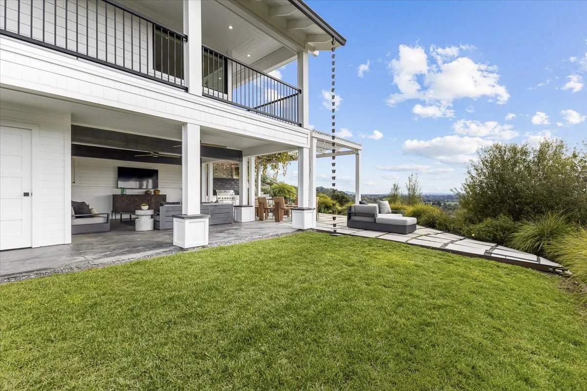 Backyard patio with outdoor furniture, a TV, a dining area, and scenic view with blue sky and trees.