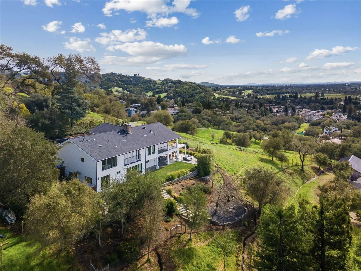 A large white house with a gray roof situated on a lush green hillside, surrounded by trees and open land, with a view of rolling hills and a distant town under a partly cloudy sky.