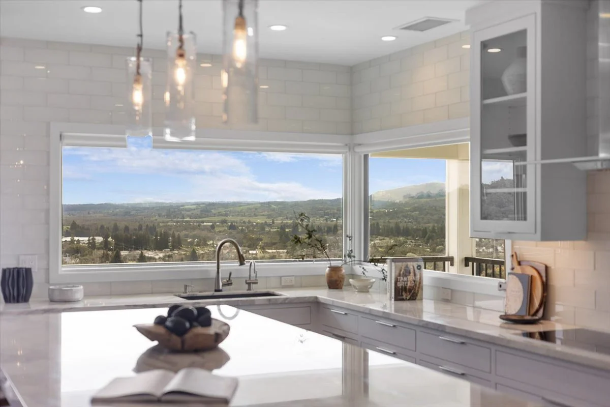 Modern kitchen with large windows showcasing a scenic landscape of hills and fields, white cabinets, and a marble countertop.