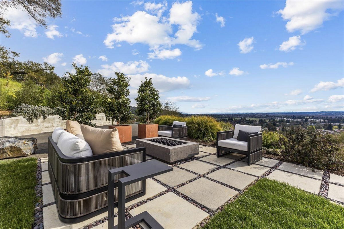 Outdoor patio with modern furniture, including a sectional sofa, armchairs, small tables, and a fire pit, overlooking a scenic landscape with trees and a blue sky with clouds.