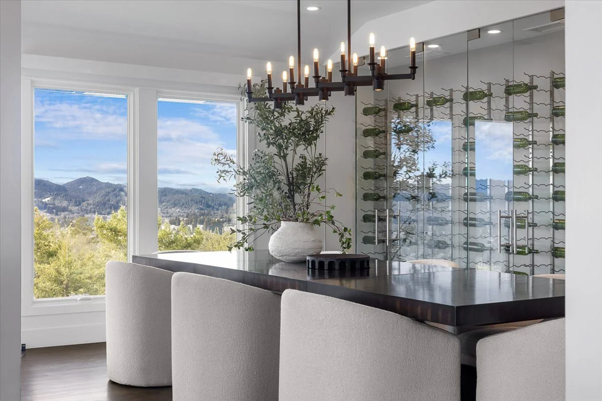 Modern dining room with a dark wood table, white upholstered chairs, a plant in a white pot, a chandelier with candle-style lights, and large windows showing a scenic mountain view.
