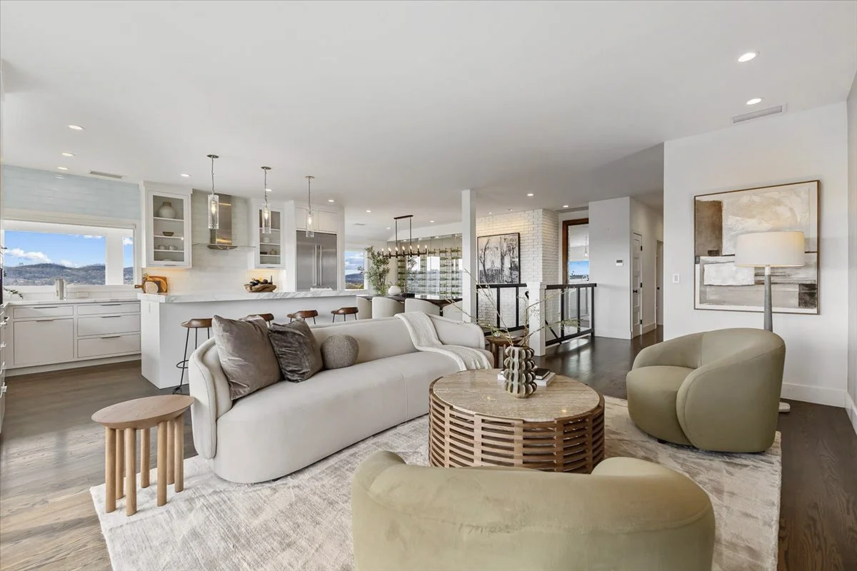 Open-concept living room and kitchen with modern decor, neutral colors, large windows showing an outdoor view, and minimalist furniture including a white sofa, green armchair, and round coffee table.