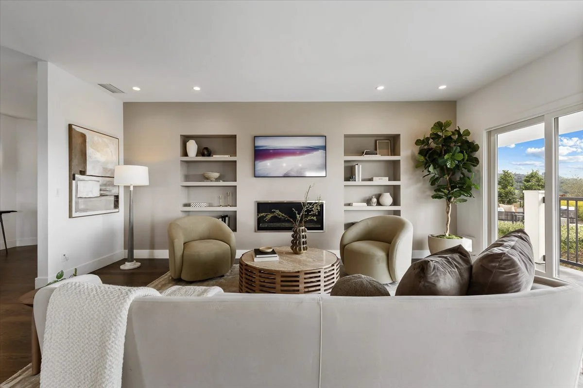 Modern living room with two beige armchairs, a white sofa with a throw blanket, a round wooden coffee table, a tall floor lamp, a large potted plant near sliding glass doors, and built-in wall shelves with decorative items and a TV.