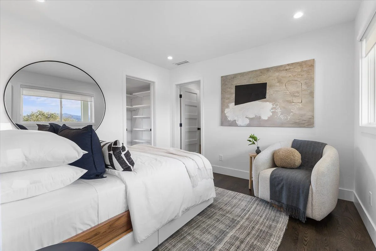 Bright bedroom with white walls, a bed with white bedding and navy and patterned pillows, a round mirror reflecting a window, a small armchair with a throw blanket and pillow, a side table with a plant, a painting on the wall, and a window with natur