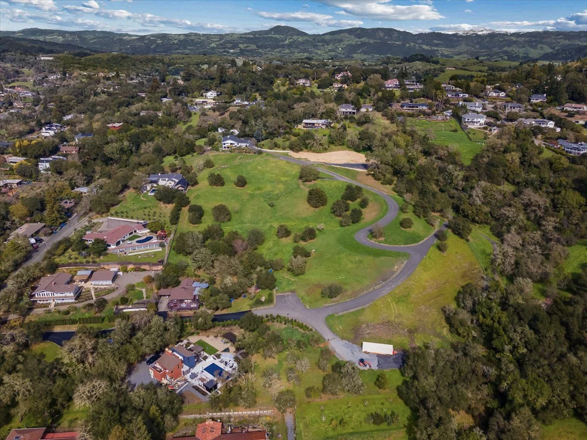 Aerial view of a residential neighborhood with houses, streets, and a large green park with trees and a winding road, set in a hilly landscape with mountains in the background.
