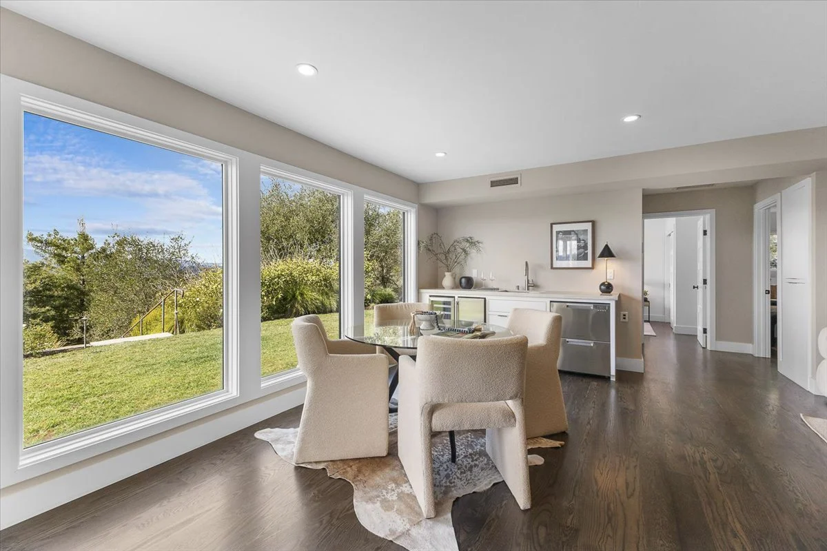 Dining area with large windows showing a green yard and blue sky, beige chairs around a glass table, and a sideboard with decor in a modern, bright home.