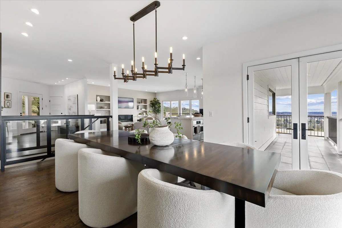 Interior view of a modern living space with a wooden dining table, white upholstered chairs, a chandelier, and large windows revealing an outdoor balcony with a view.