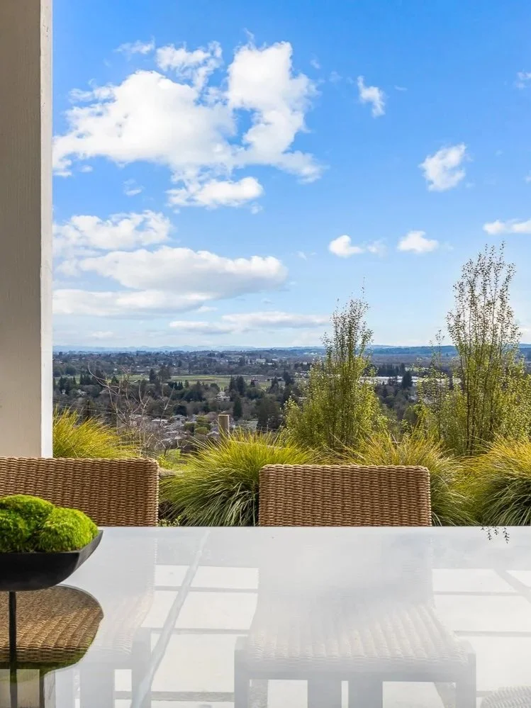 View of a balcony or patio with a glass table and wicker chairs, overlooking a landscape with trees and a cityscape under a partly cloudy blue sky.