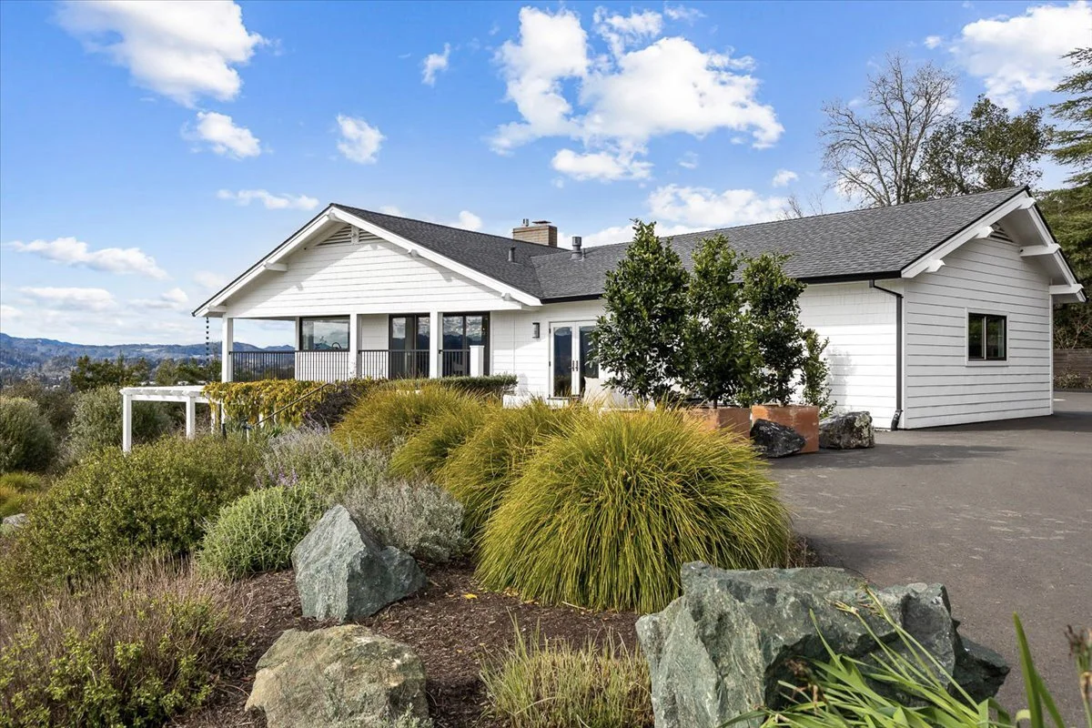 A white house with black roof, surrounded by landscaped garden with rocks, plants, and trees under a blue sky with scattered clouds.
