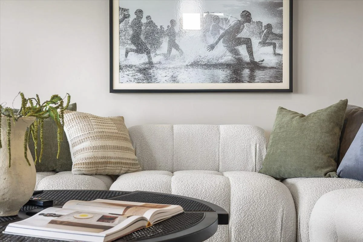 Living room with a white textured sofa, decorative pillows, a black coffee table with an open magazine, a potted plant, and a large black-and-white photograph of children playing in the rain hanging on the wall.