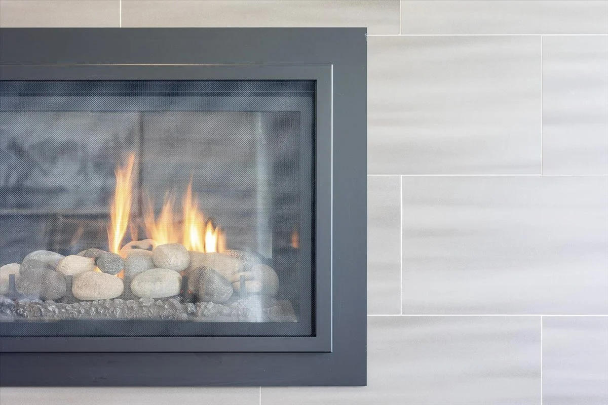 A modern fireplace with a glass front, showing flames and rocks inside, set against a light-colored tiled wall.