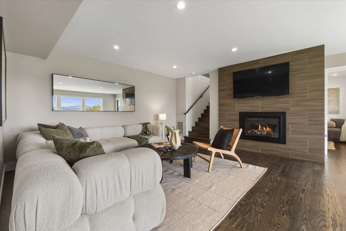 Living room with a large white sectional sofa, a black round coffee table with a vase, a lounge chair, a fireplace with a mounted TV above, and a staircase in the background.
