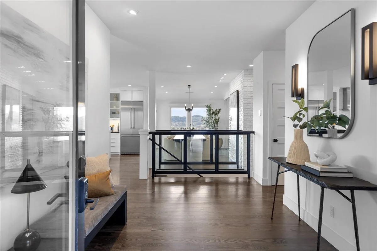 Modern open-concept living area with a view of a kitchen and dining space, featuring white walls, wooden floors, a black railing, and minimal decor.