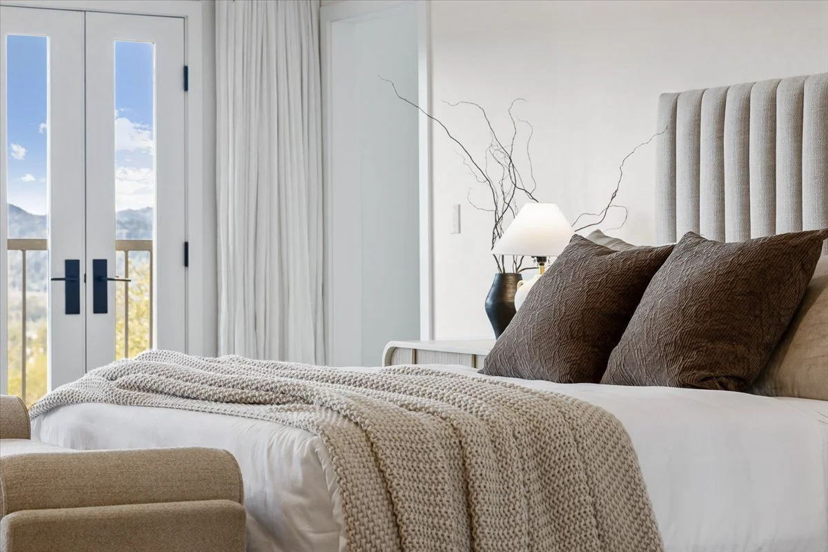 Bedroom with a large bed, brown pillows, beige knitted blanket, bedside table with a black vase, white lamp, and decorative branches, with closed double glass doors leading to a balcony with mountains and blue sky in the background.