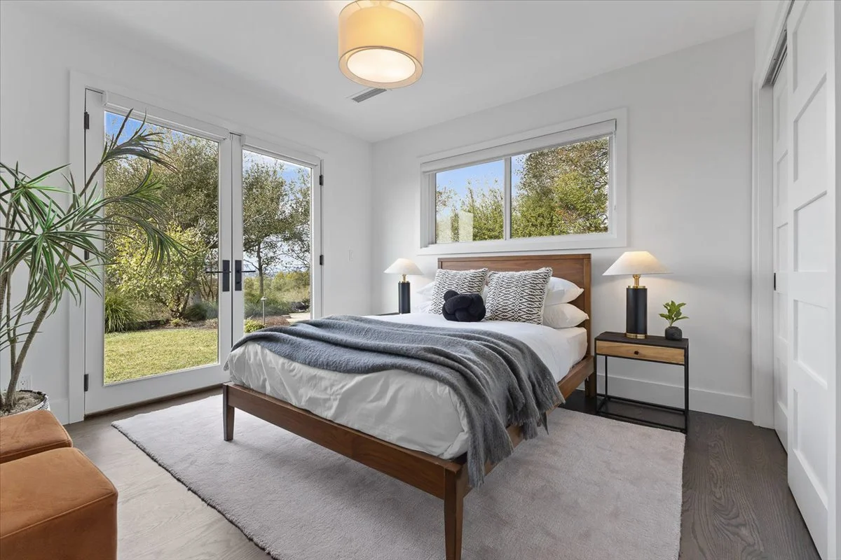 A bedroom with a large bed, nightstands with lamps, a sliding glass door, and a window showing trees outside.