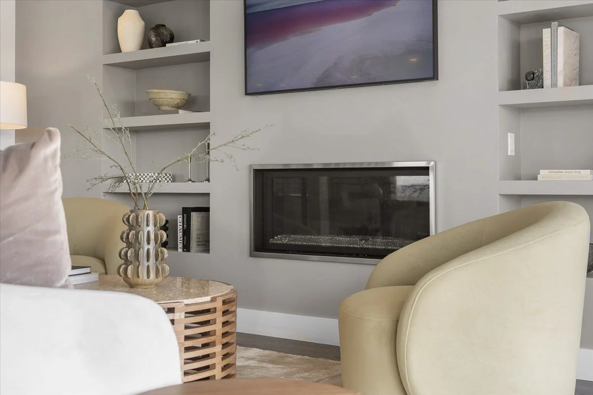 Living room with built-in wall shelves, a modern fireplace, mounted TV, beige armchair, a round coffee table with a decorative vase, and books on shelves.