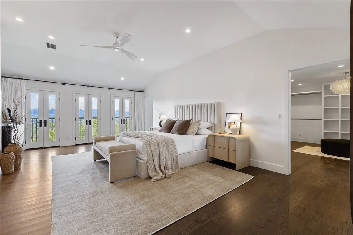A bedroom with a large bed, side tables with lamps, a beige armchair at the foot of the bed, a rug, and a view of mountains through glass doors.