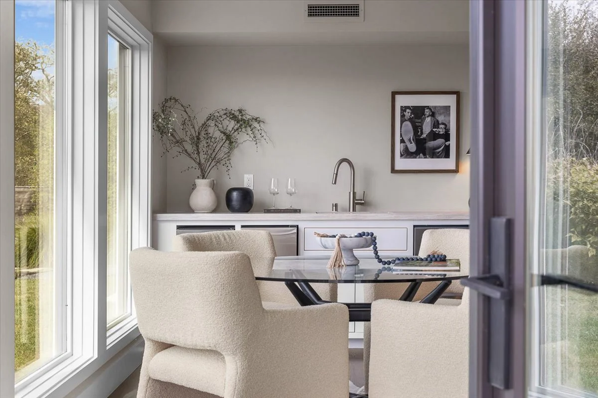 Dining area with four cream-colored chairs around a glass table, and a kitchenette with a sink and minimalist decor in the background, seen through a partially open door or window.
