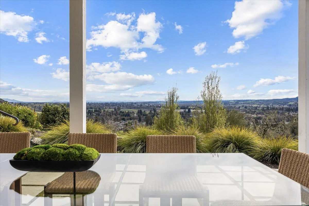 Outdoor patio with a glass table and wicker chairs, overlooking a scenic landscape with trees, hills, and a partly cloudy sky.