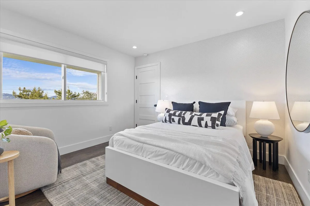 A modern bedroom with a large window showing trees and blue sky outside, a white bed with blue pillows and a black and white throw pillow, a nightstand with a lamp, and a circular mirror on the wall.