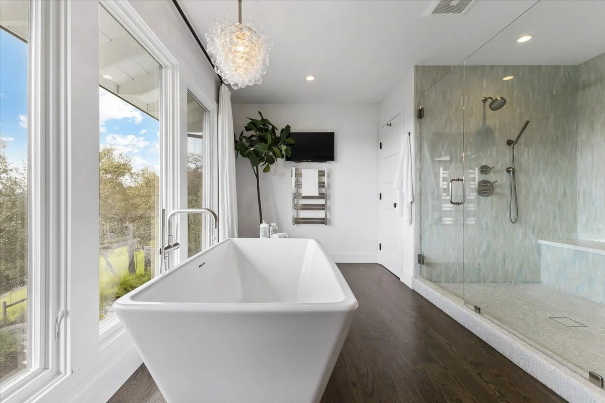 Modern bathroom with a freestanding white bathtub, large windows with curtains showing a green outdoor landscape, a glass-enclosed shower, a small TV, a towel rack, and a potted plant.