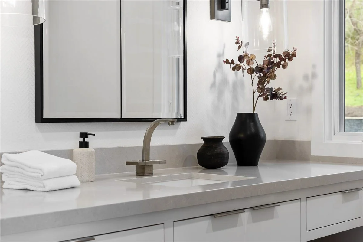 Bathroom vanity with a mirror, faucet, three folded towels, a soap dispenser, a black vase with dried leaves, a smaller black vase, and a window showing greenery outside.
