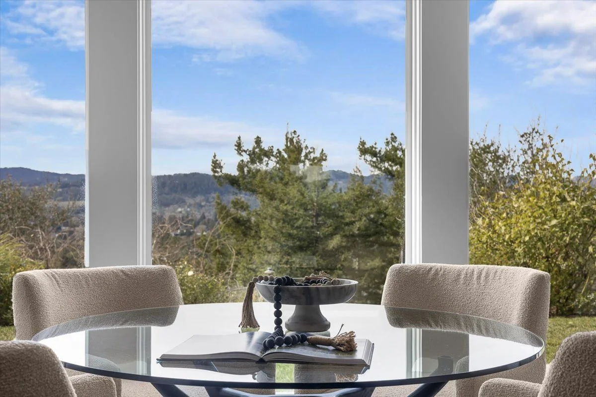 A modern dining area with a round glass table, three beige upholstered chairs, a windowsill with a scenic view of trees and distant hills, a bowl of beads, and an open book.