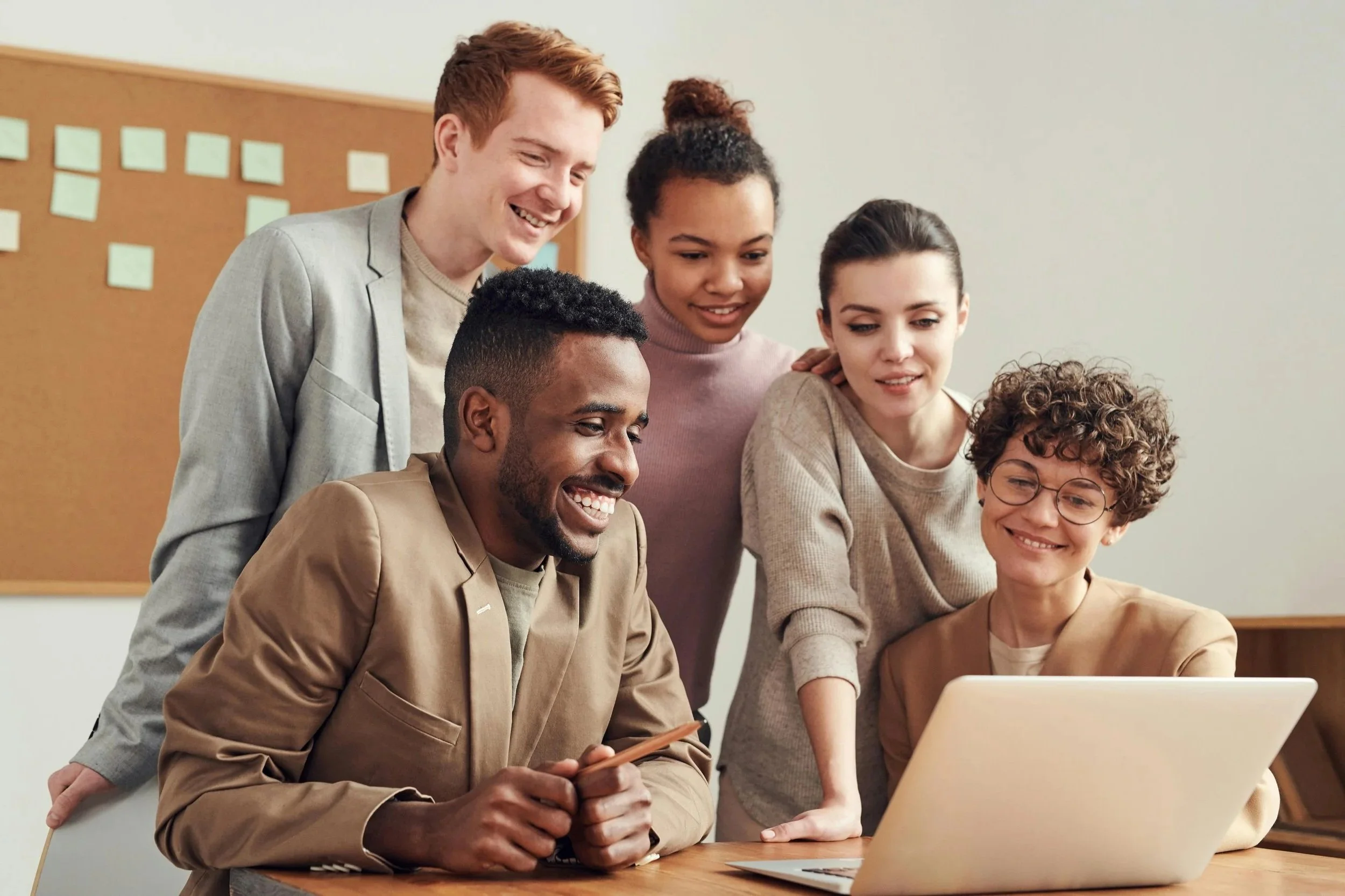 Group of six diverse young adults gathered around a laptop, smiling and engaged in a discussion in a bright office.