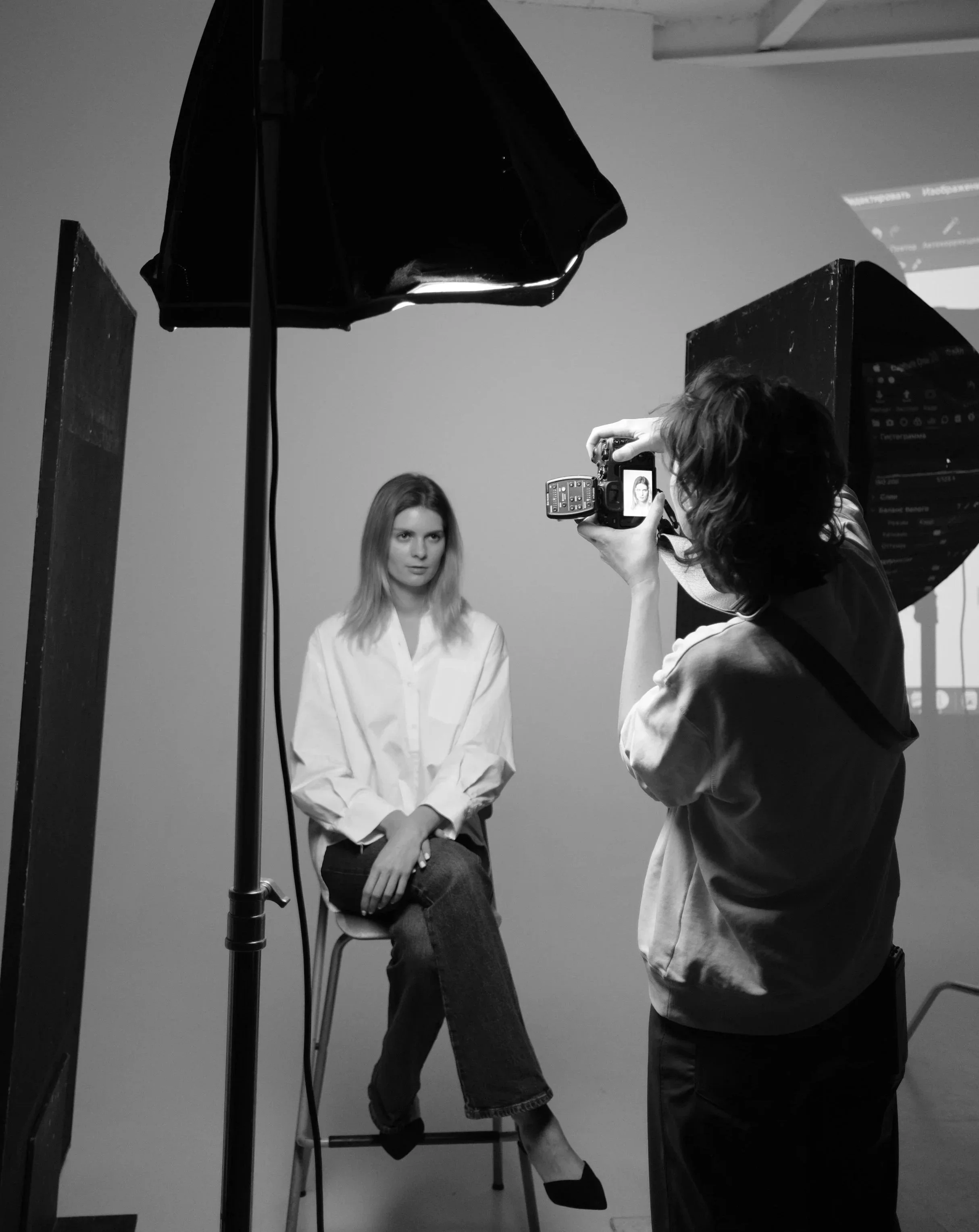 A photographer taking a portrait of a woman sitting on a stool in a studio with lighting equipment and a plain background.