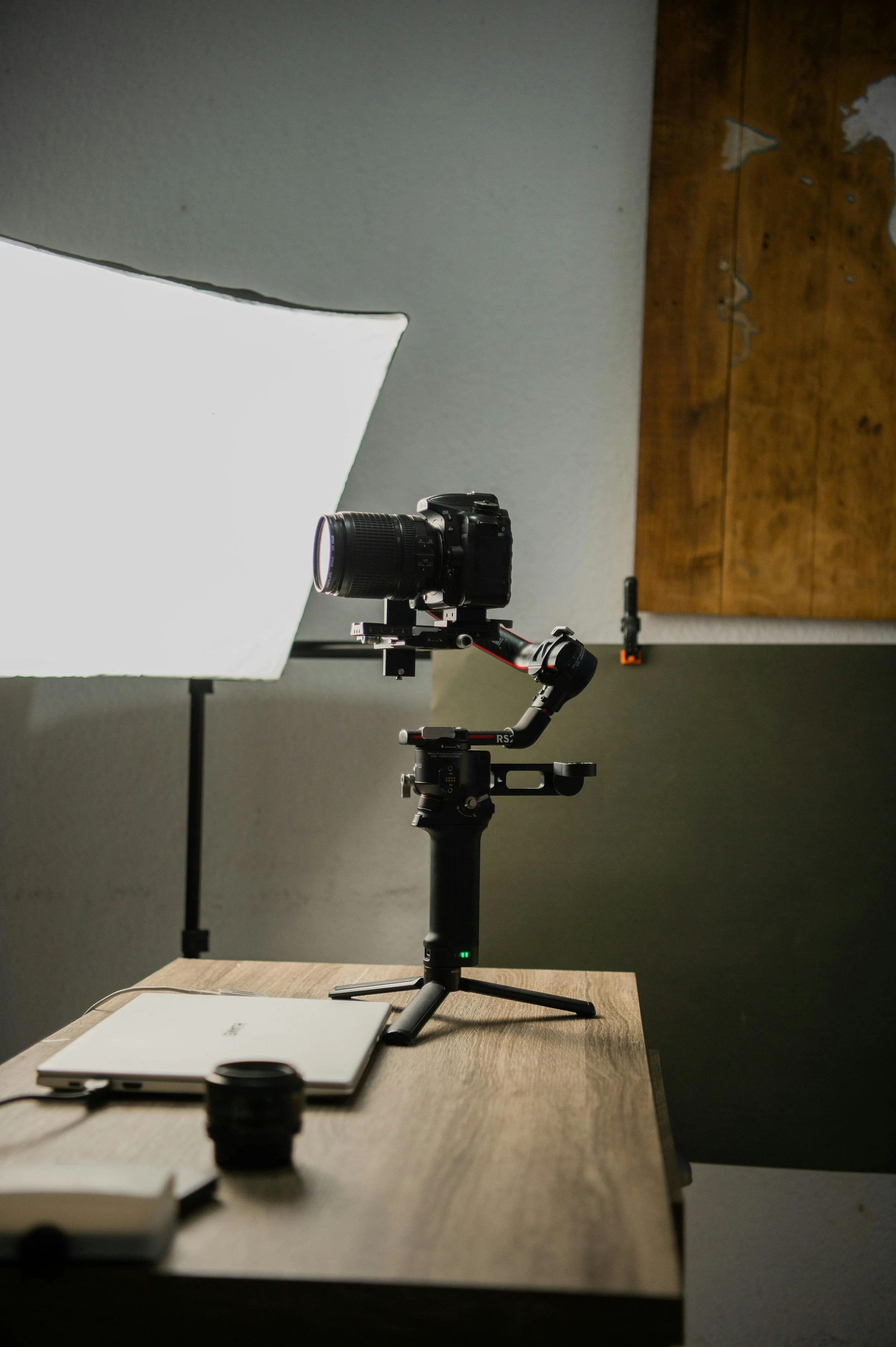A camera mounted on a handheld stabilizer on a wooden table, with a laptop, a camera lens, and a wireless remote control nearby, set up for filming or photography in front of a large softbox light.