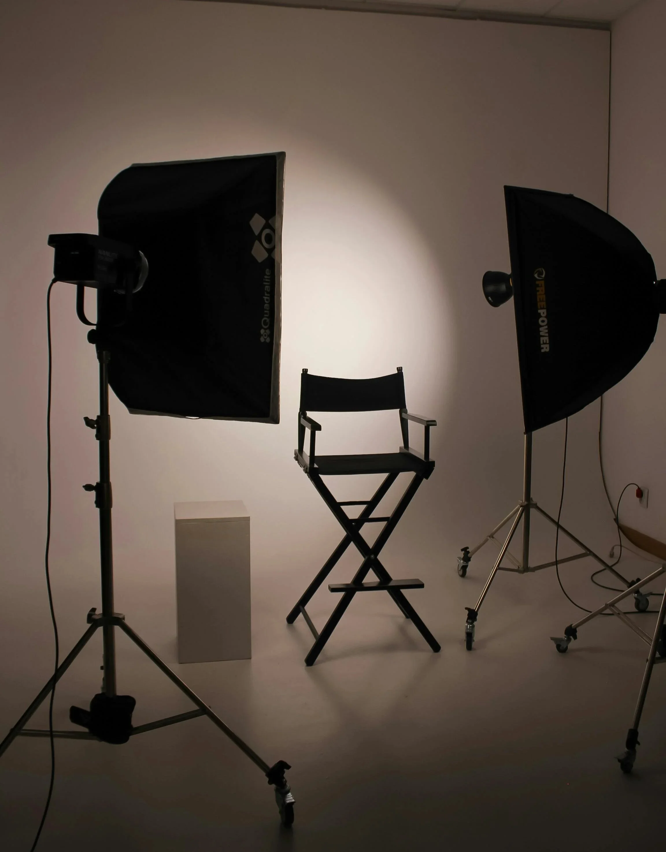 Photo of a photography studio setup with two softbox lights, a director's chair, and a white pedestal against a plain white background.
