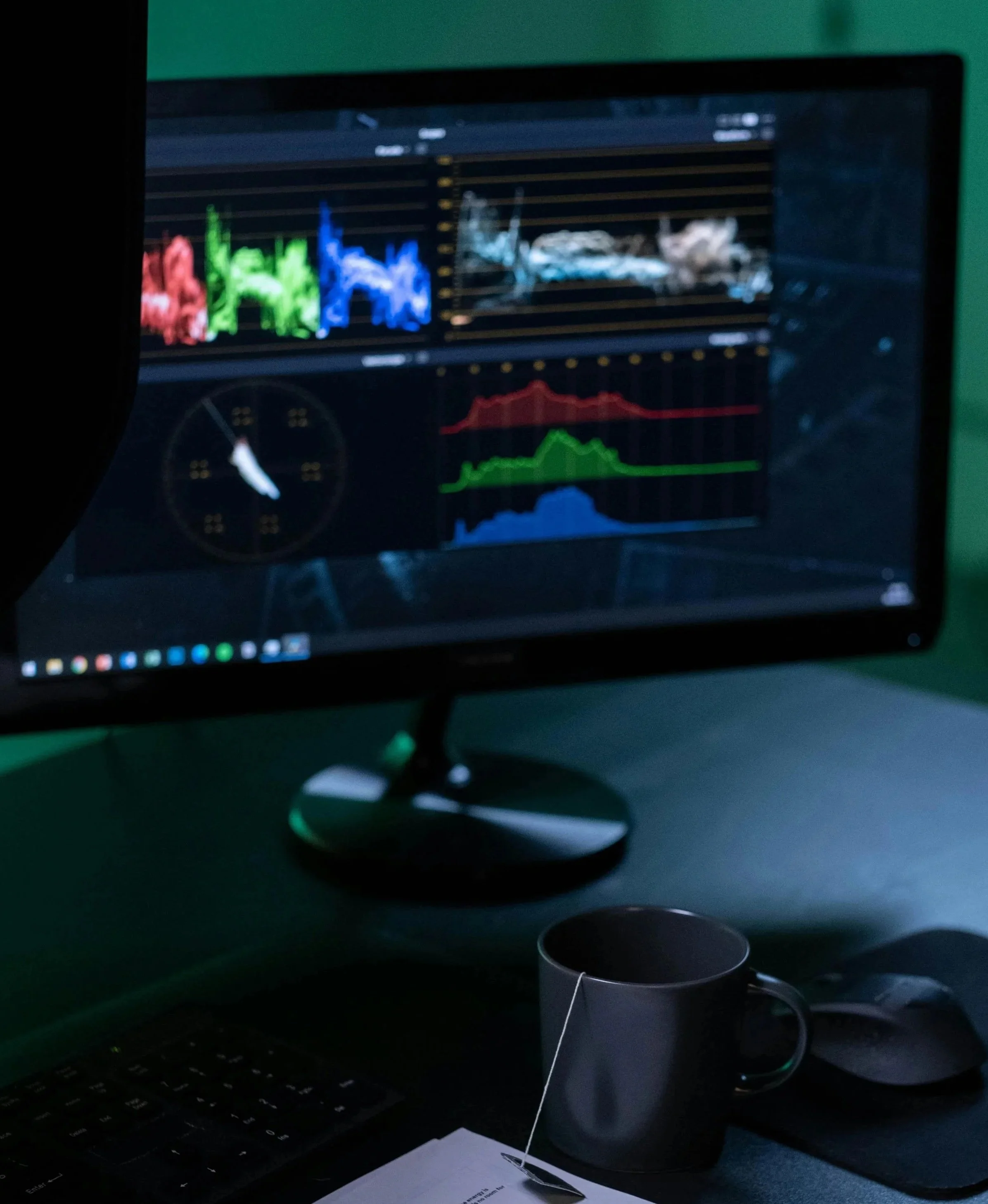 Computer monitor displaying various colorful charts and graphs; a black mug and a computer mouse on a dark desk with a document and pen nearby.
