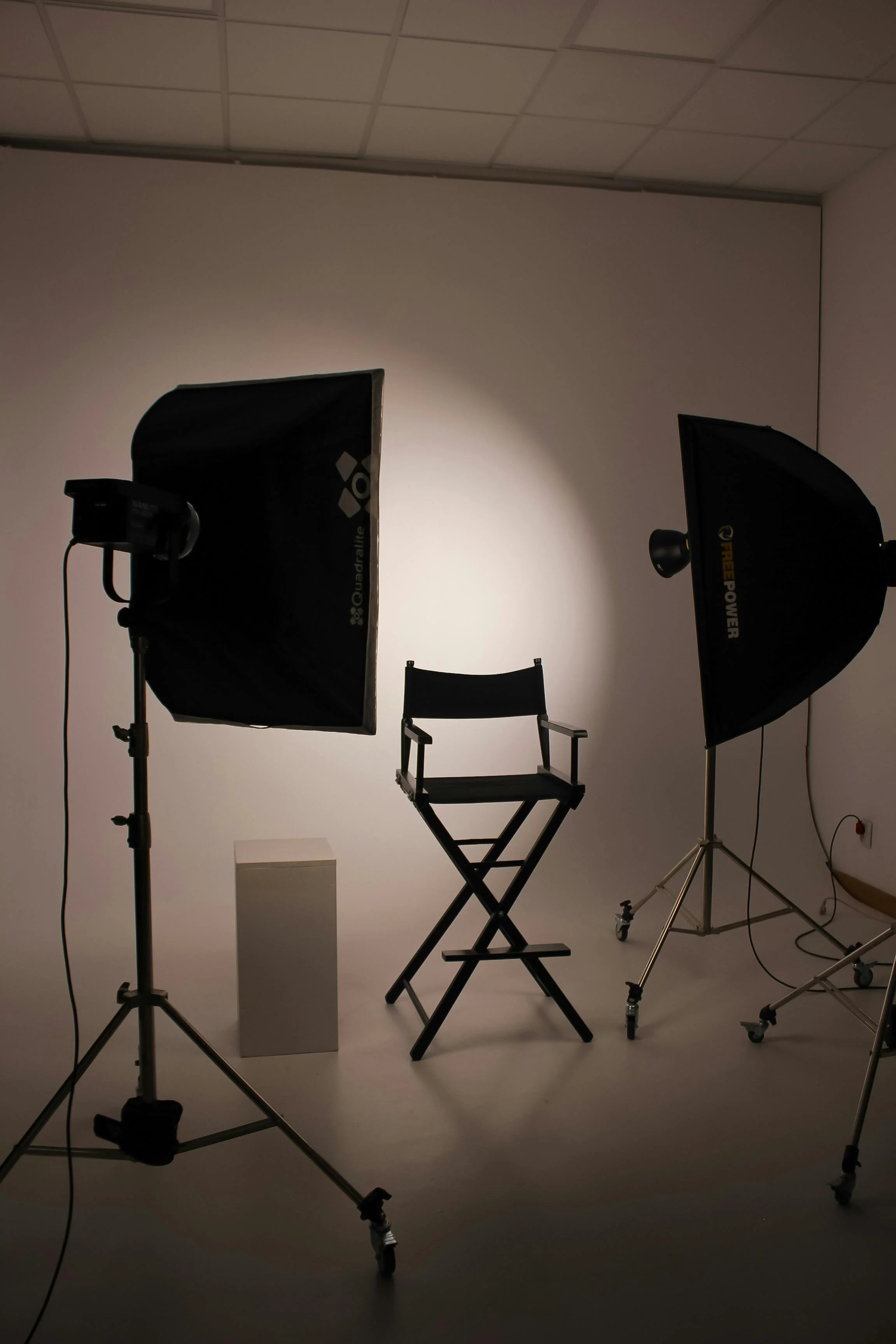 Photography studio with two large studio lights directed towards a blank white backdrop, a director's chair, and a small white pedestal.