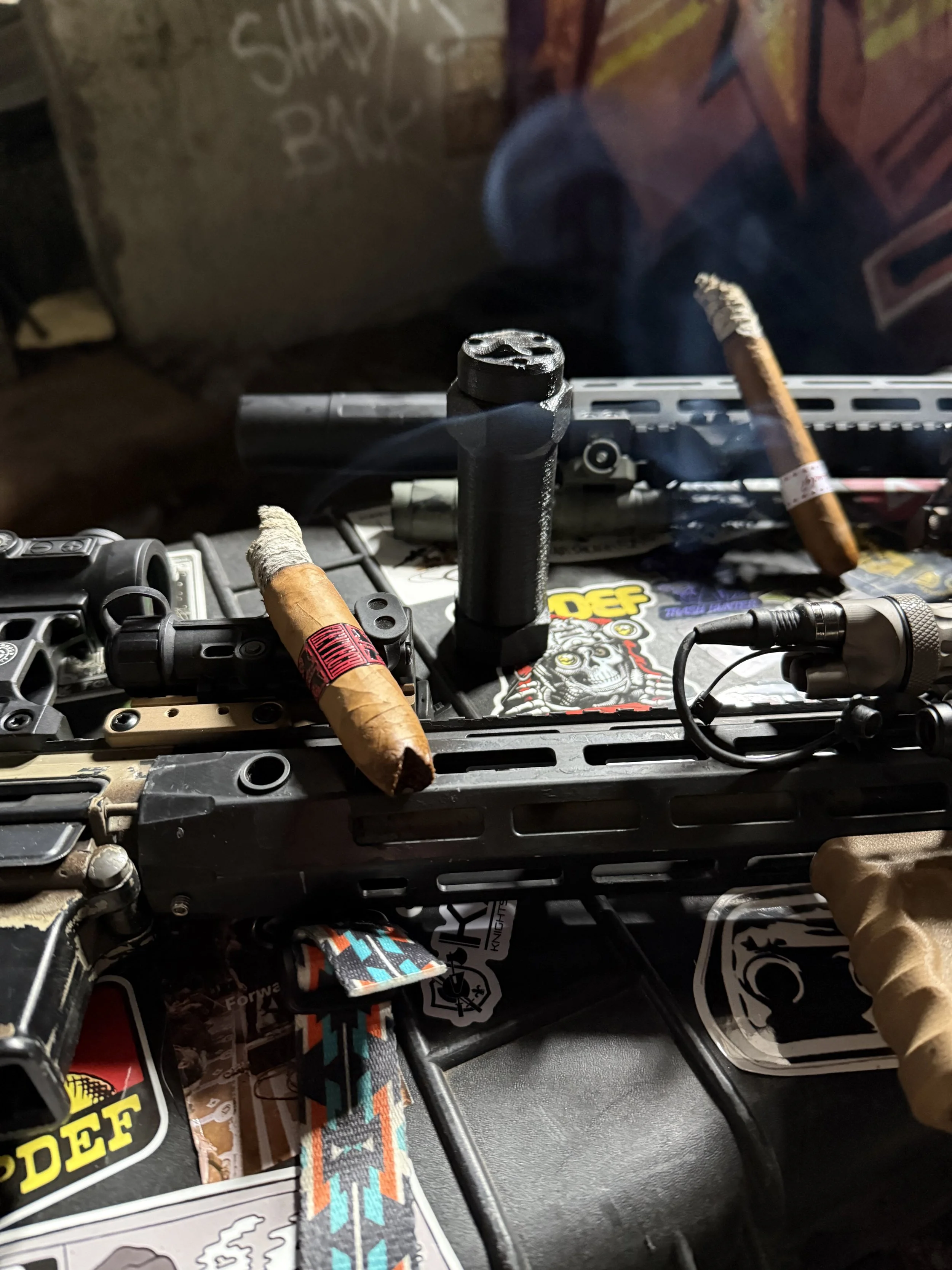 Close-up of a firearm, partially concealed by two cigars, with a black cylindrical suppressor attached, resting on a surface decorated with stickers and patches.