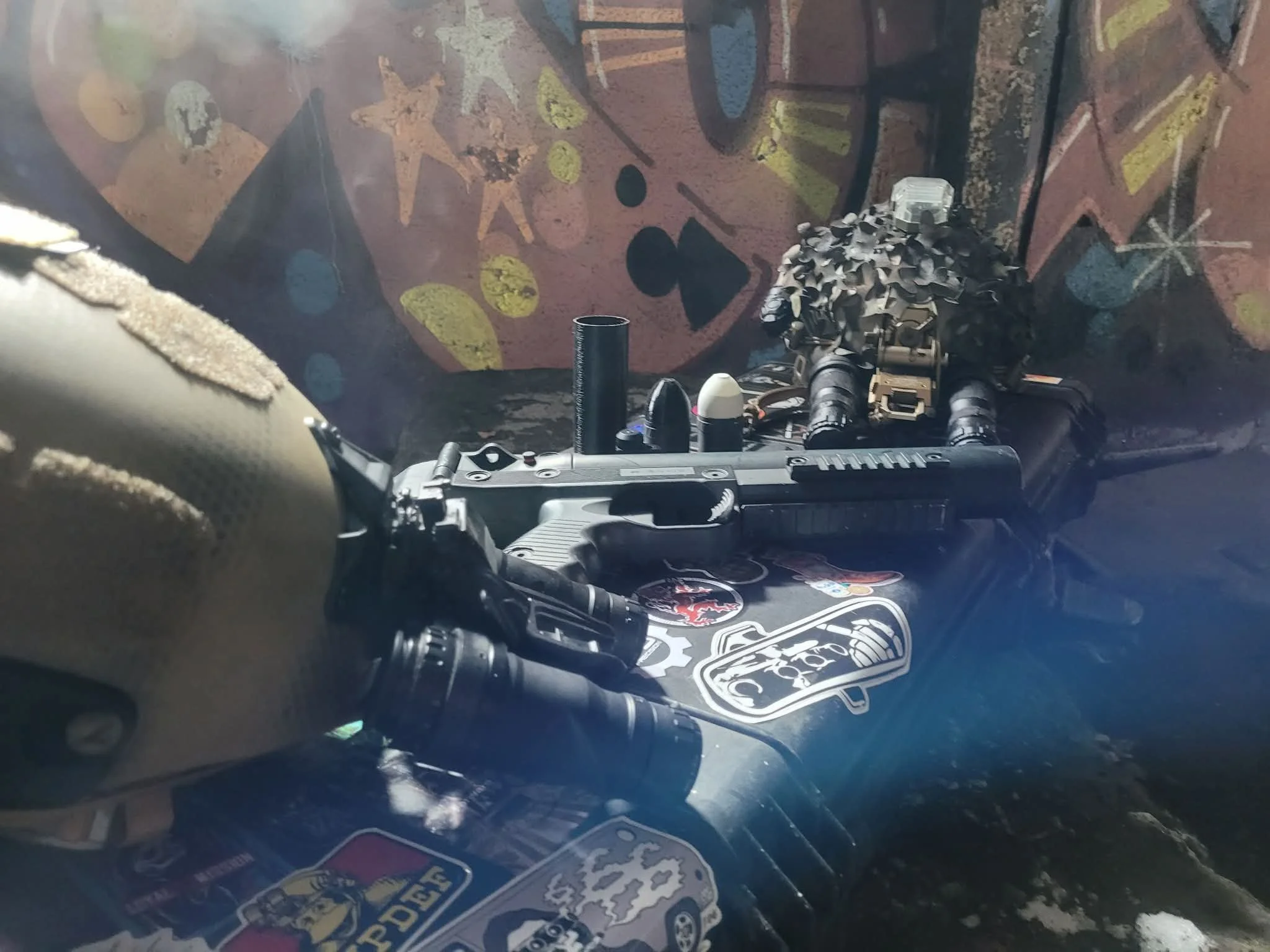 A collection of military-style gear including a helmet, a handgun, and a camouflage-covered object on a surface with stickers and graffiti.
