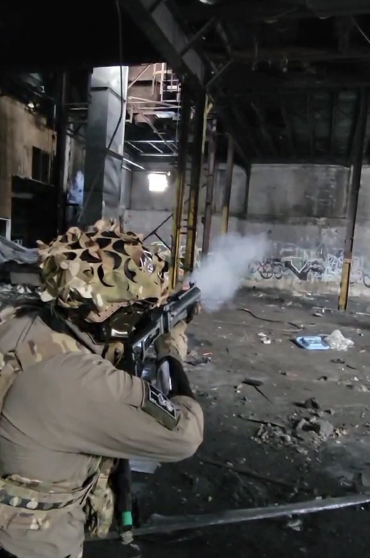 Soldier in beige camouflage uniform firing a gun inside an abandoned industrial building with graffiti on the walls.
