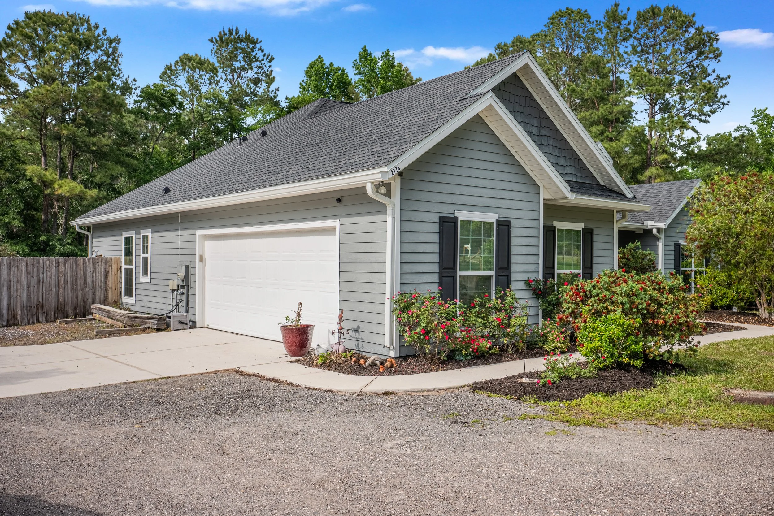 A house with gray siding, black shutters, and a two-car garage surrounded by a small garden with bushes and flowers, with a gravel driveway and a wooden fence on the left side, set against a background of green trees and a blue sky.