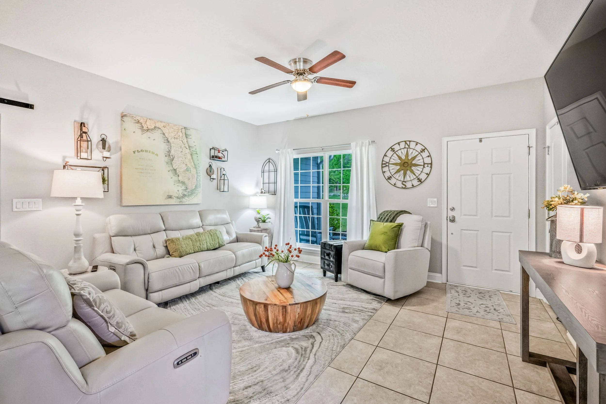 Bright living room with white walls, white sofa, armchair, coffee table, ceiling fan, wall art, and large window with white curtains.