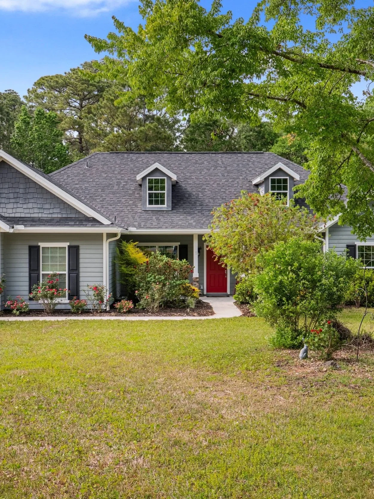 A house with a gray exterior, black shutters, a red front door, surrounded by green trees and shrubs, with a grass lawn in the foreground and a clear blue sky.
