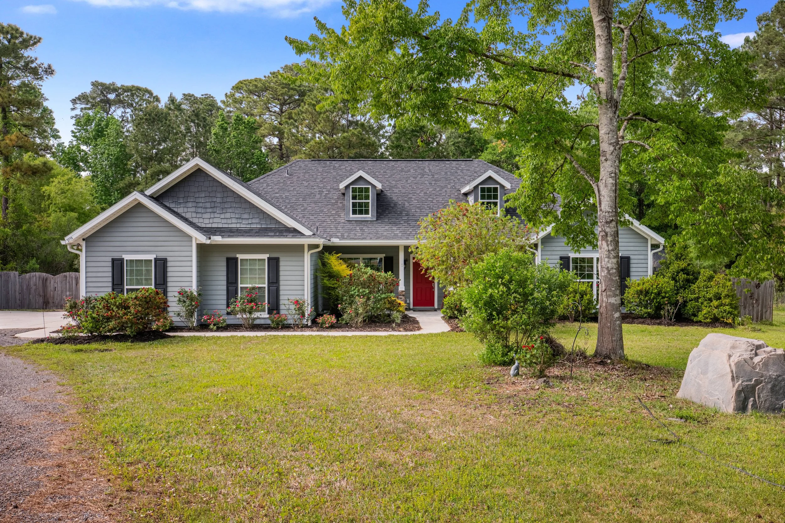 A light blue house with white trim and a red door, surrounded by green trees, shrubs, and a lawn, with a clear blue sky overhead.