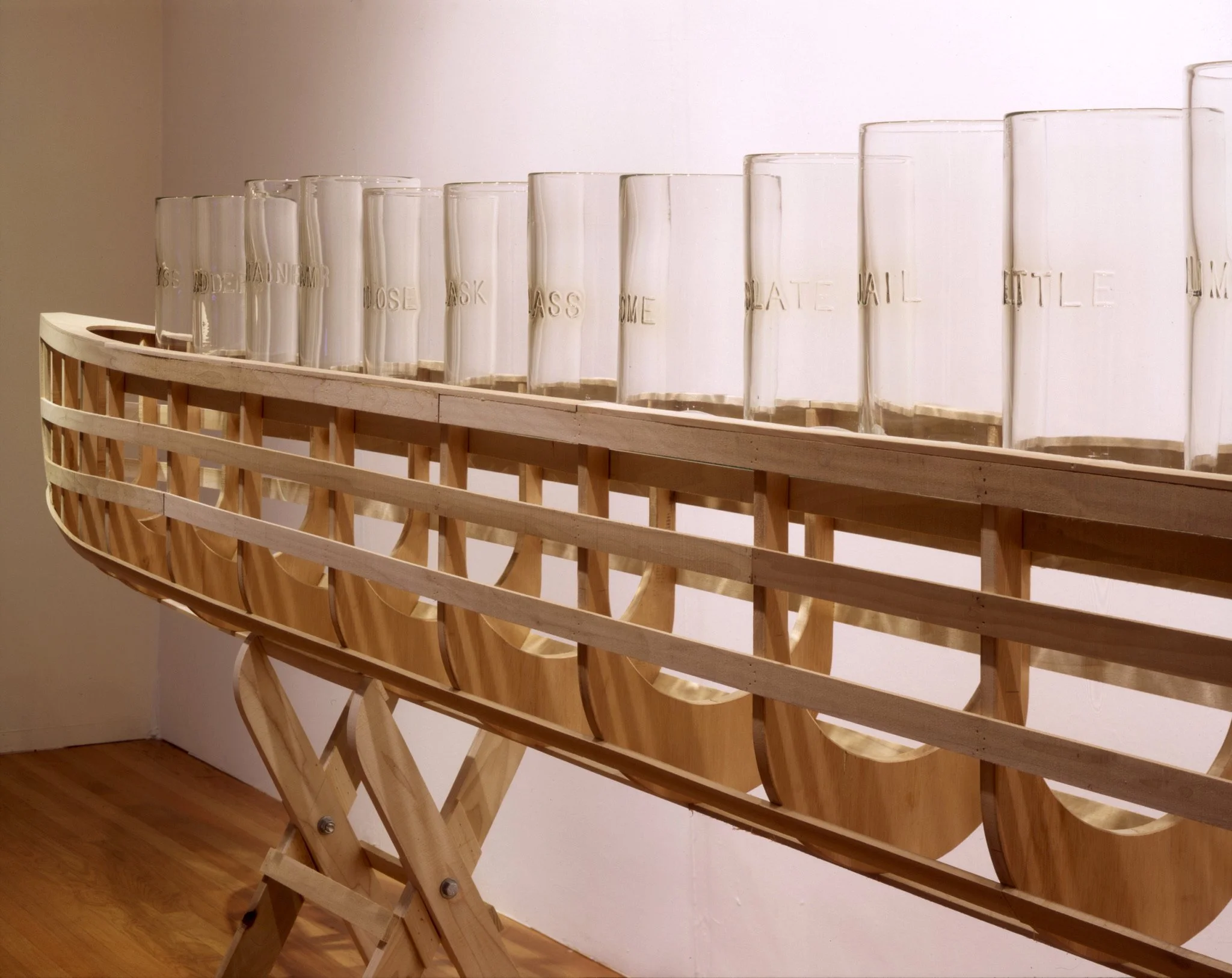 A wooden ship model with transparent glass bottles on top, each labeled with phrases like 'use model,' 'rose ask,' 'passport,' 'home,' and 'date mail,' displayed against a plain wall with a wooden floor.