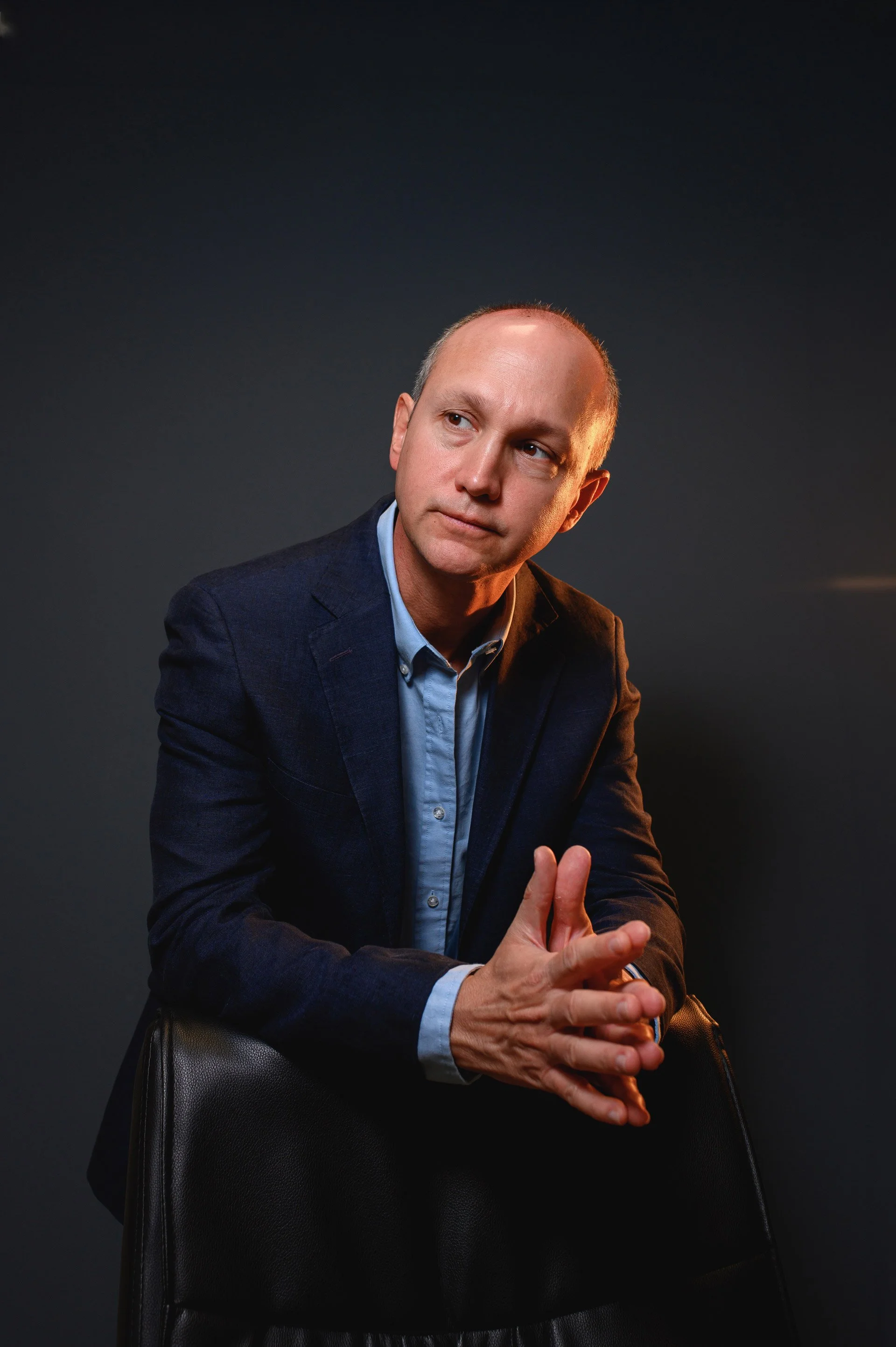 Hombre de aspecto pensativo, vestido con saco azul y camisa, apoyado en una silla negra, con fondo oscuro.